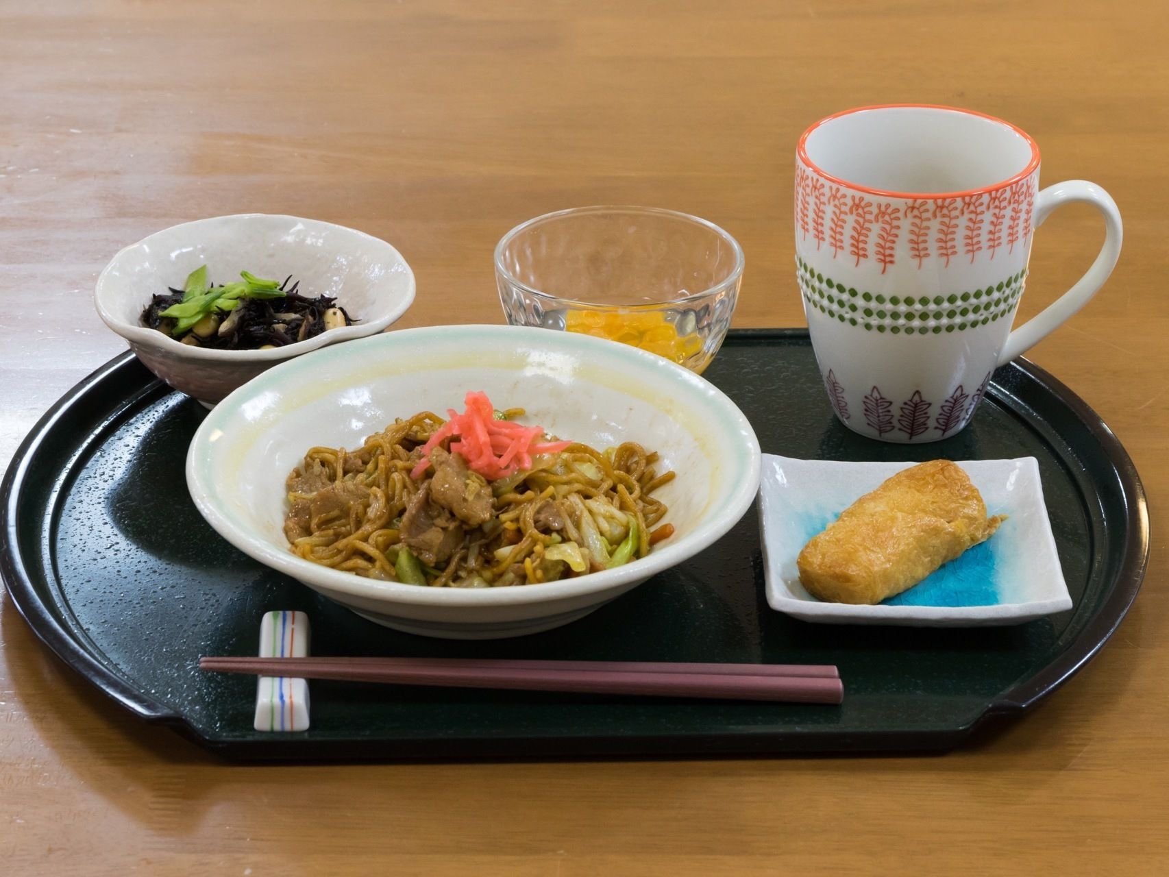 やきそばに稲荷ずし、ひじきの煮物とオレンジがそれぞれ皿に分けて盛られている。黒いお盆の上に、きれいに並んでいる。