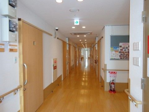 廊下の壁には居室のドアが並んでいる。天井に複数の丸い照明が設置されており、手前のスペースには消火器が置かれている。