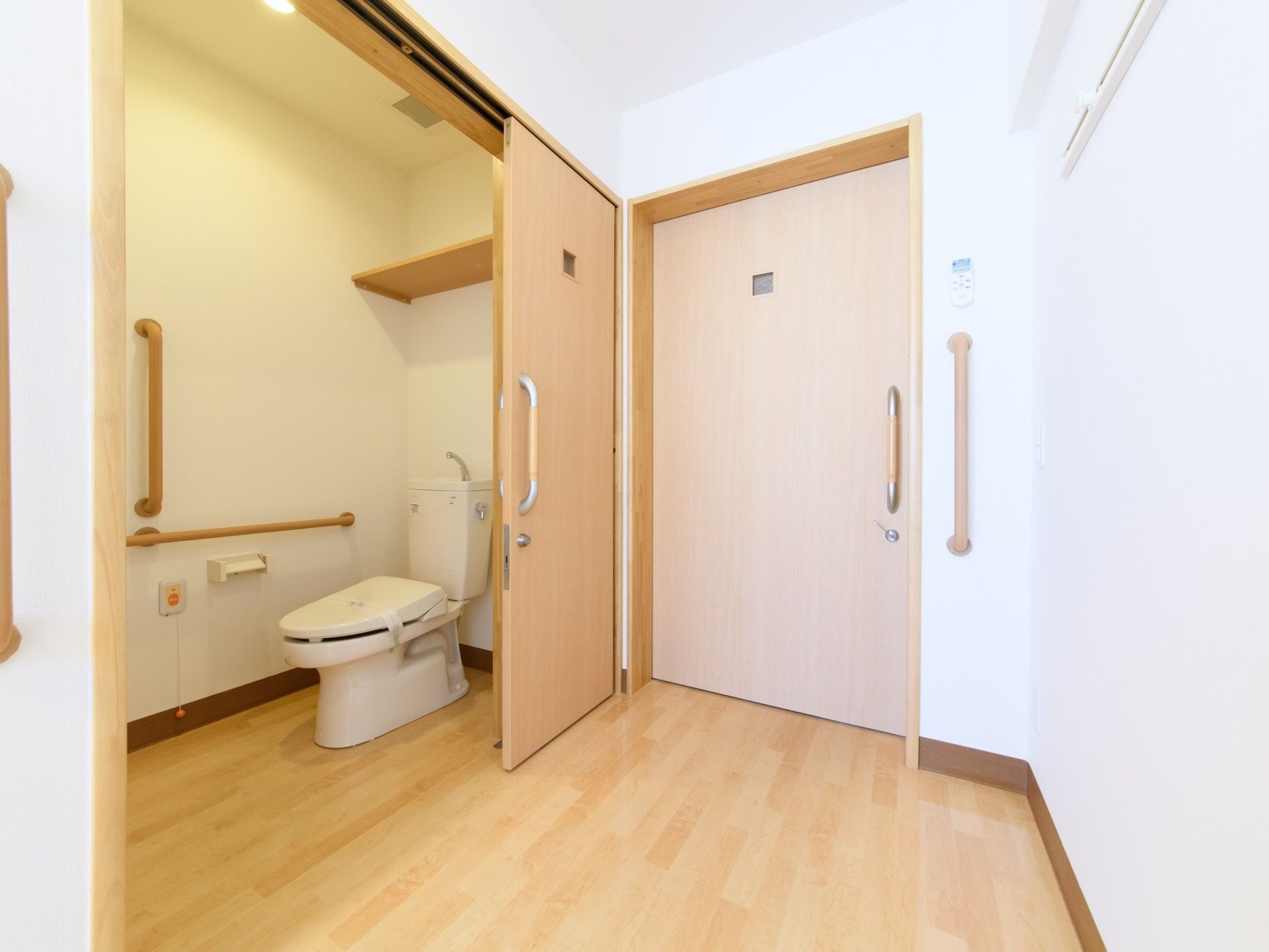 トイレの出入り口には、木目調デザインの引き戸が設置されている。奥の壁には、オレンジ色の緊急呼出ボタンや手すりがある。