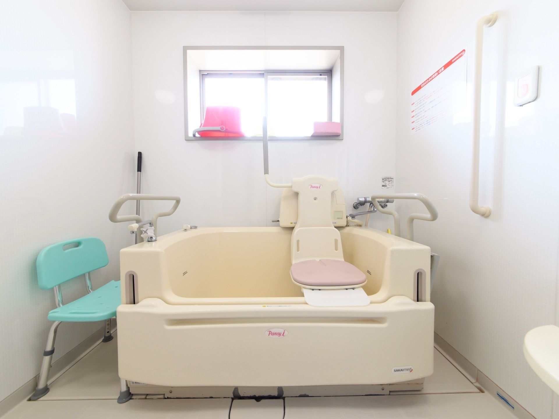 バスタブの右端に、座浴用の機械が設置されている。正面の壁には窓があり、ピンク色のバケツが窓枠のスペースに置いてある。