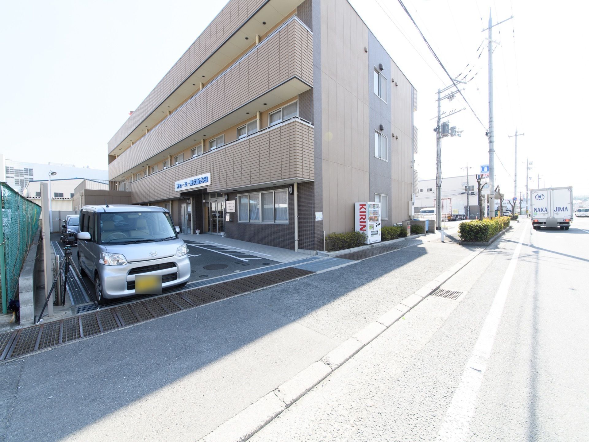 地上３階建ての施設を、斜め方向から写したものである。左側には駐車場があり、シルバーの軽自動車が置いてある。