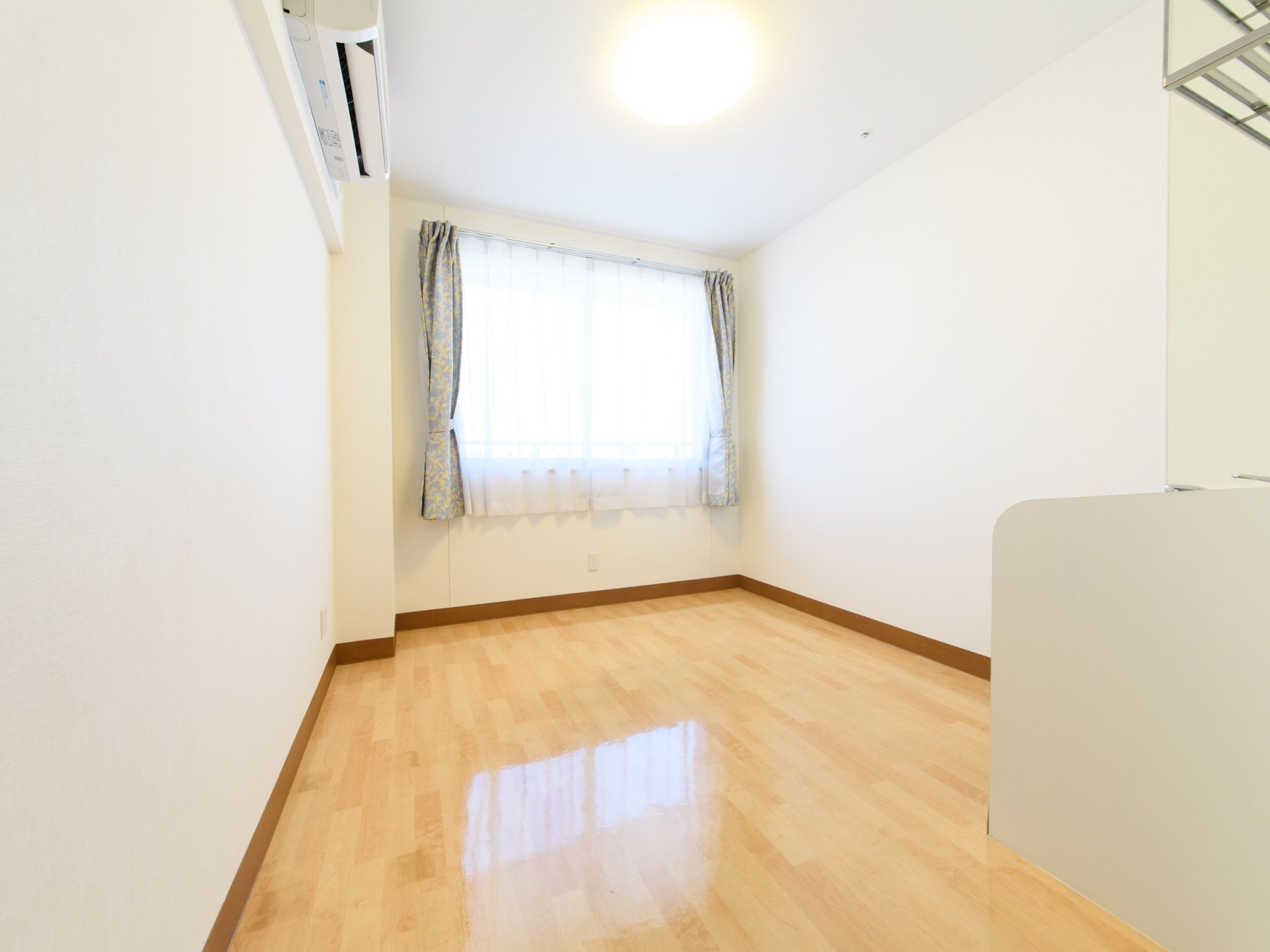 出入口のほうから見た居室の様子である。部屋の奥に窓があり、明るい光が差し込んでいる。天井近くにエアコンが設置されている。