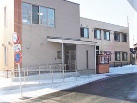 ブラウン系の2階建ての建物。玄関部分には屋根があり、階段とスロープも付き、道路に面している。