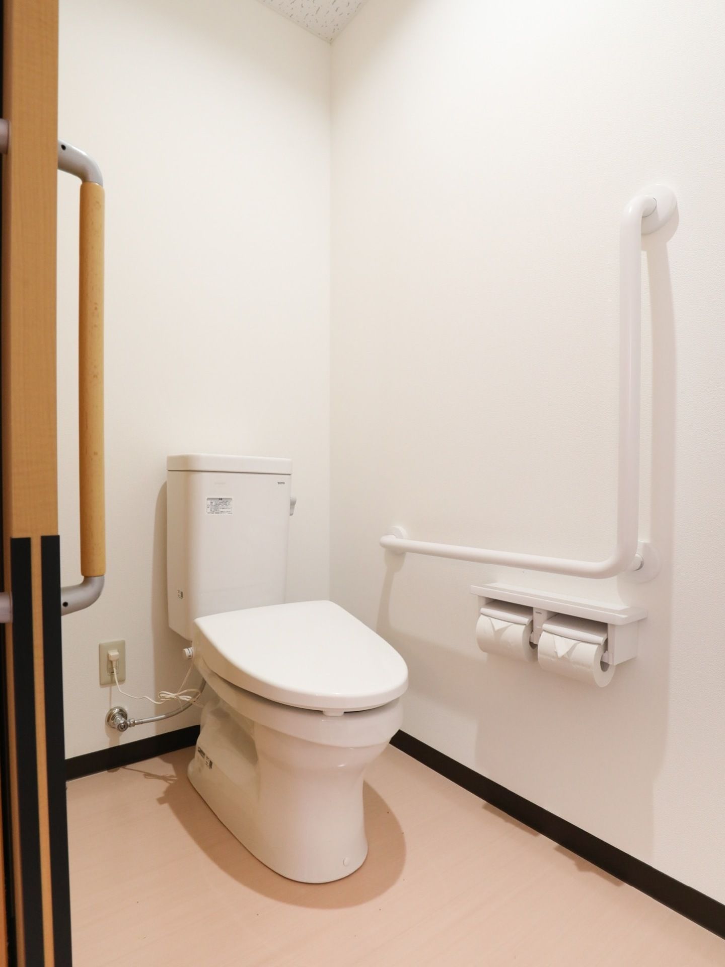 壁から電源がとられたタンク式の洋式トイレが設置されている。壁に手すりと二連タイプのペーパーホルダーが付いている。