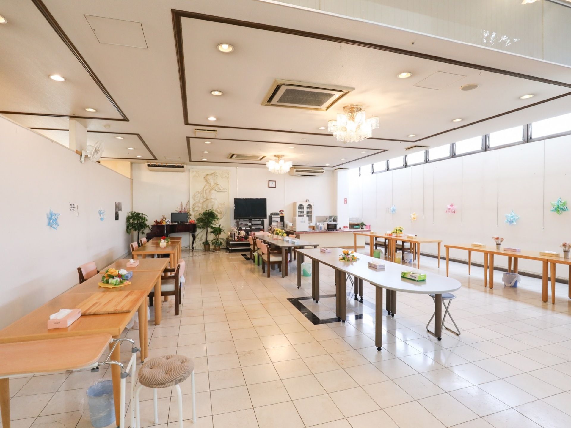天井にダウンライトとシャンデリアが設置されている。部屋の中央と両側にテーブルが並べられ、壁際にはテレビと観葉植物などが置かれている。