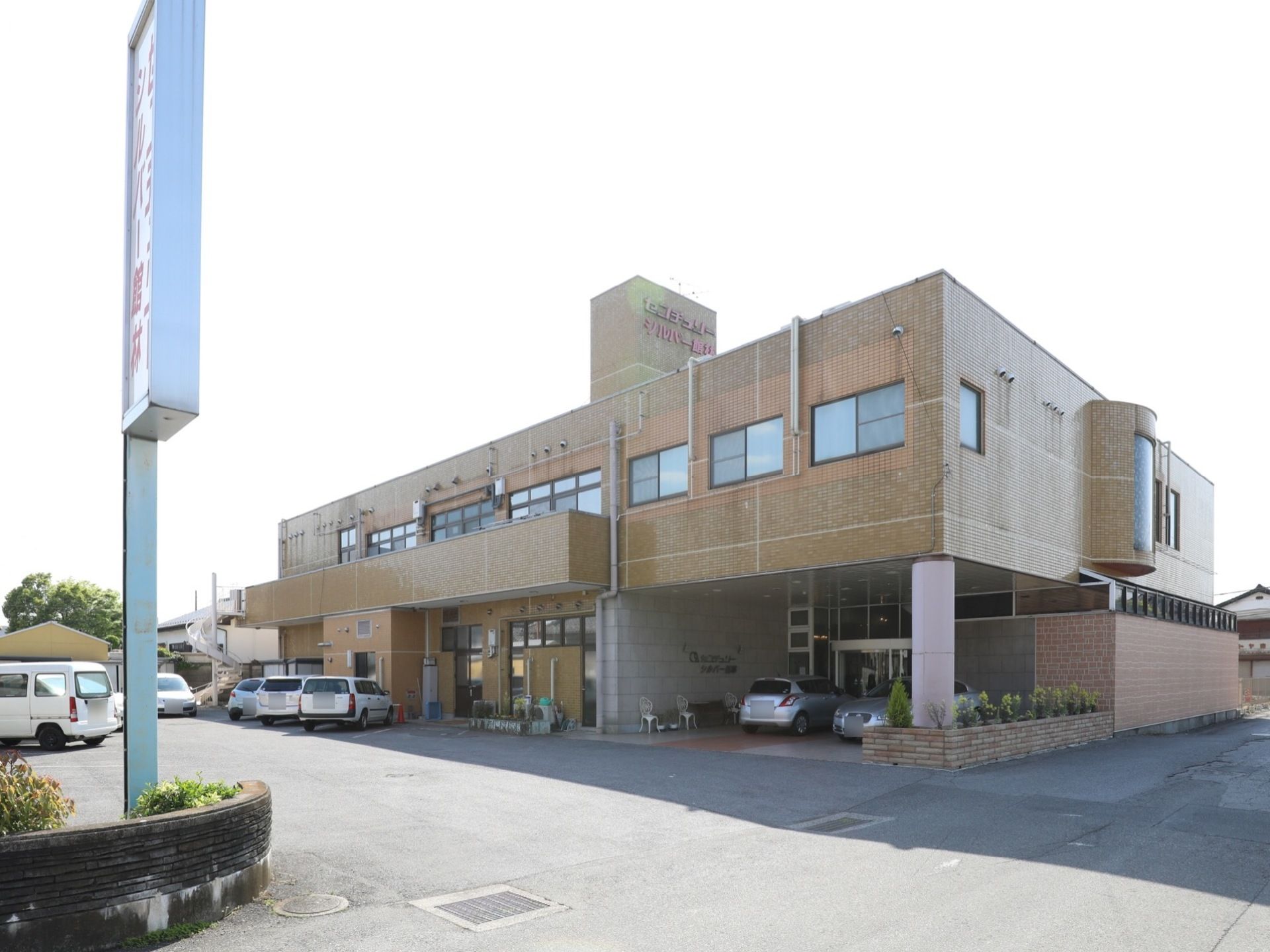 茶色いレンガ調の２階建て建物の周りに車が数台停まっている。道路からの入り口の植栽スペースに看板が立てられている。