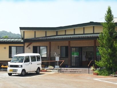 ベージュの外壁の平屋建ての建物の前には車が止まっていて、緑も多く植えられている。