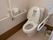 温水洗浄便座が取り付けられたトイレが中央にある。向かって右側の壁に折り畳み式の手すりがあり、伸ばされている。