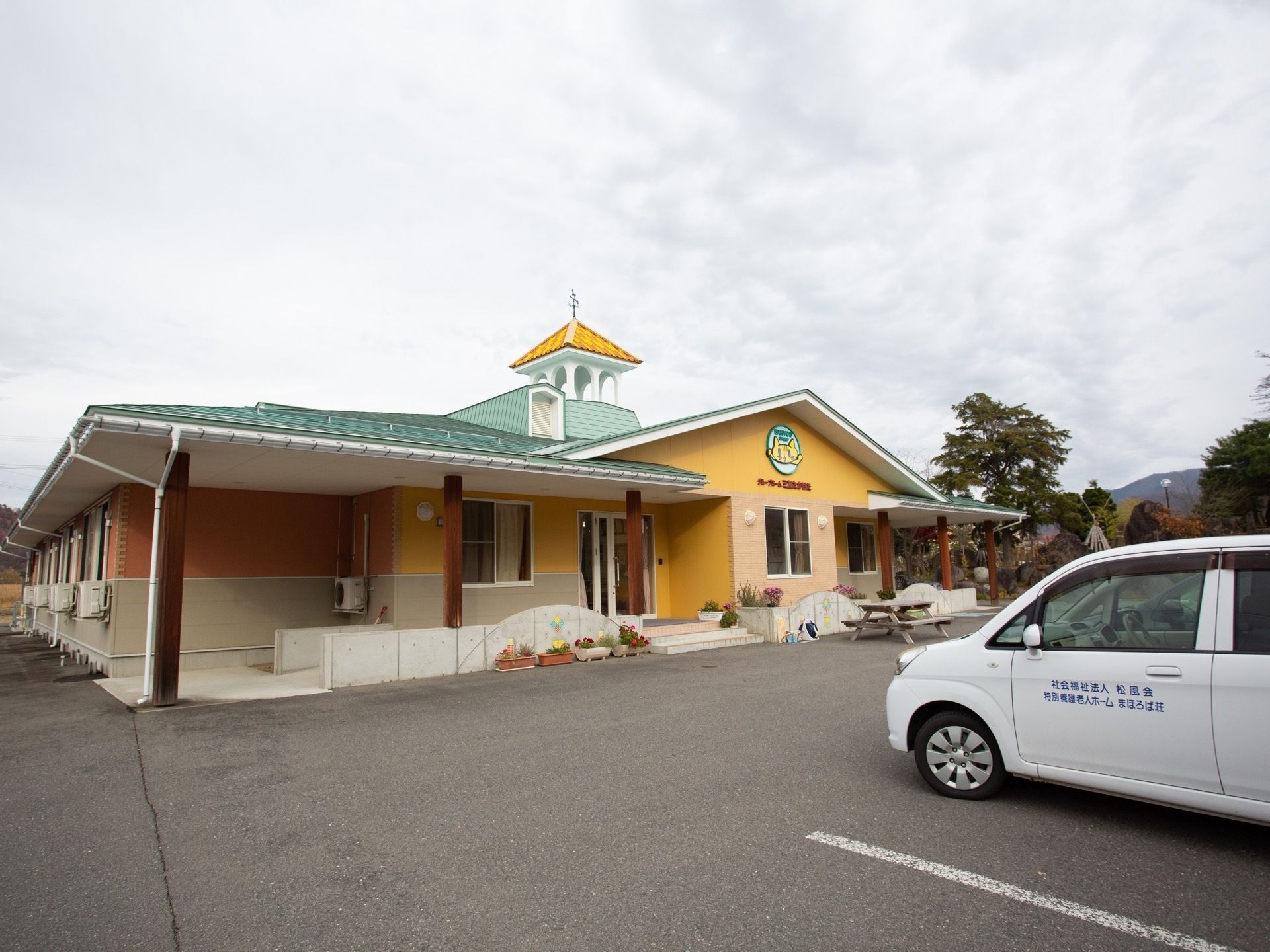 黄色い外壁に緑の屋根が乗った１階建ての建物である。施設前は駐車場になっており、白い乗用車が停まっている。