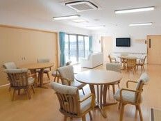 4人掛けのダイニングテーブルセットが３組置かれ、窓際には白いソファ、壁にはテレビが設置され、窓には青いカーテンが付いている。