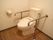 床の木目が美しい白を基調としたトイレである。温水洗浄便座の両側には、ステンレス製のしっかりとした手すりが完備されている。