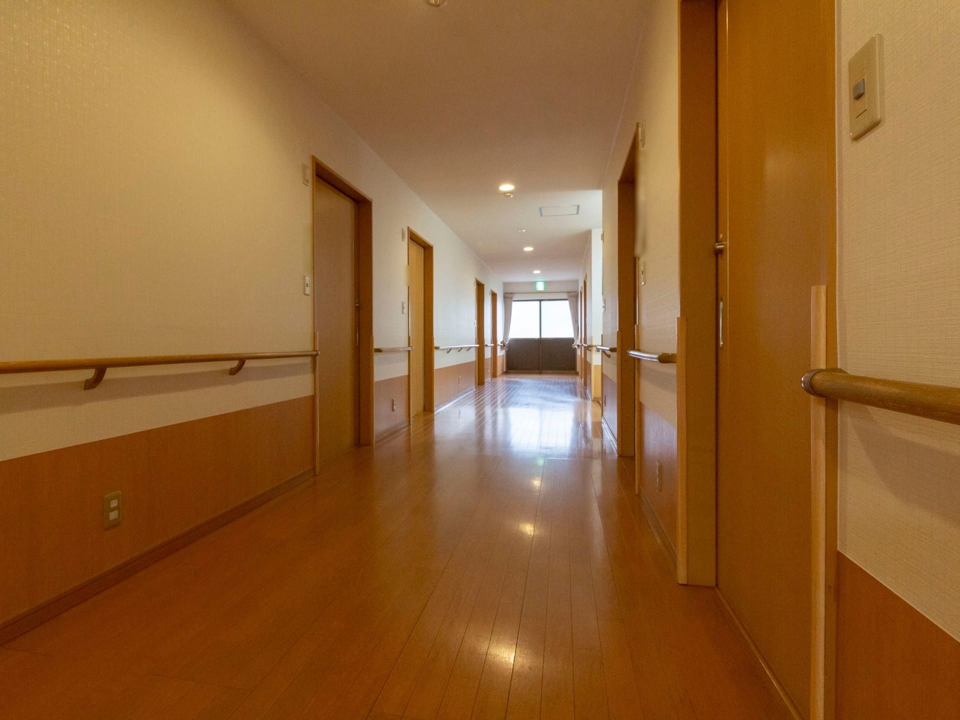 木目調のデザインが印象的な通路である。両脇には部屋が並び、壁際には手すりが設置されている。奥には非常口を知らせる標識が見える。