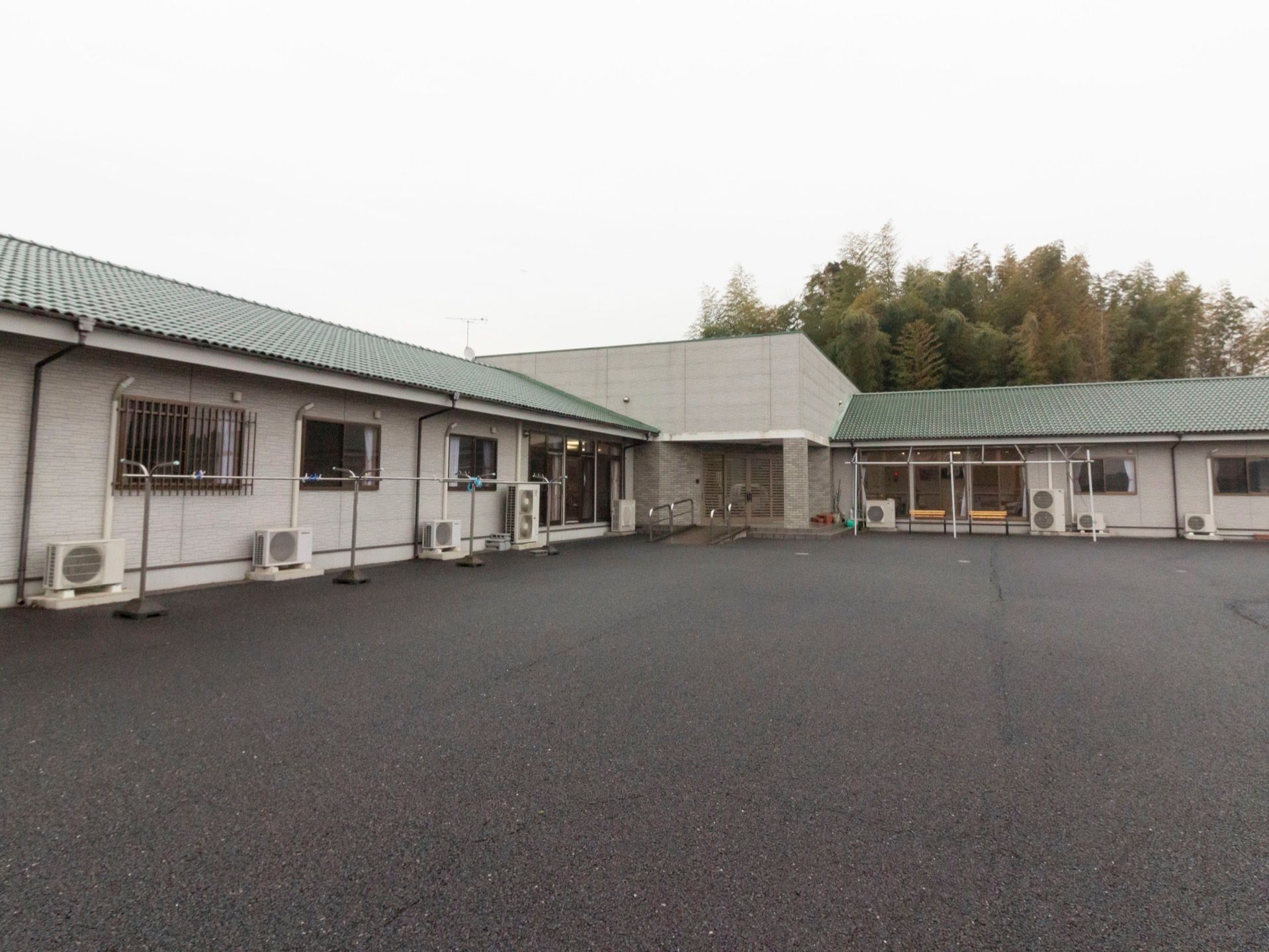 深緑色の屋根が印象的な建物が当施設である。エアコンの室外機や物干しざおが並んでいるのが見える。駐車スペースが広くとられている。