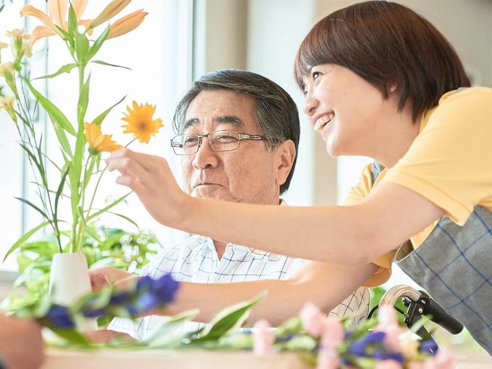 フラワーアレンジメントを楽しんでいる眼鏡をかけた高齢男性と手伝っているエプロンを付けた若い女性スタッフ。