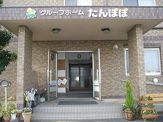 レンガ調の壁がアットホームな雰囲気の建物。玄関にはブラウンの屋根が付けられ、木や花もある。
