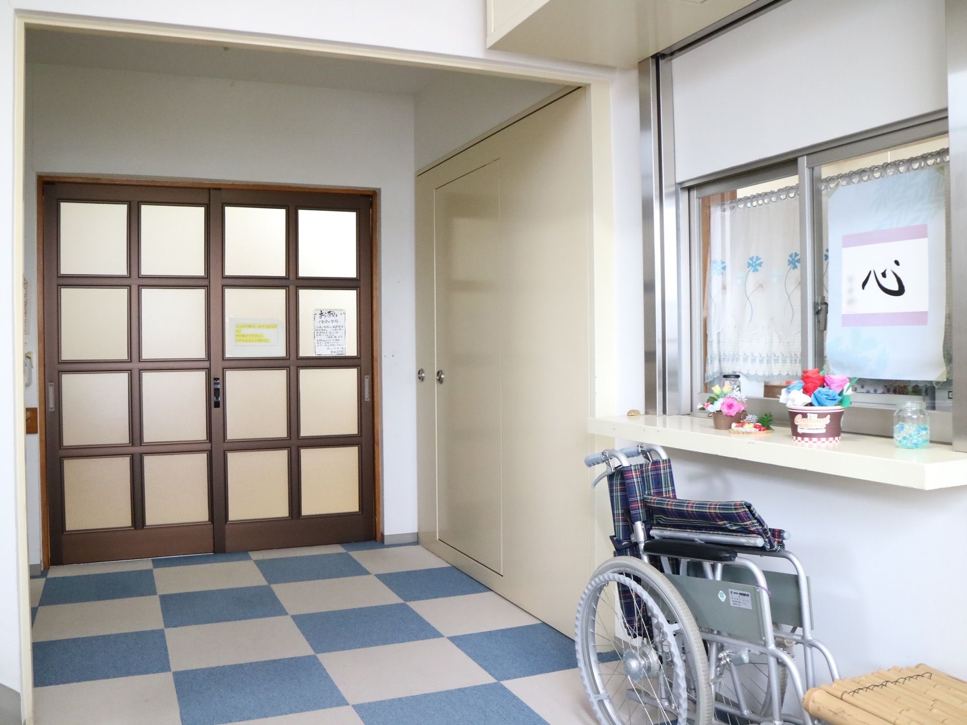 白と水色のカーペットタイルが敷かれた玄関。受付カウンターがあり、近くに車椅子とベンチが置かれている。