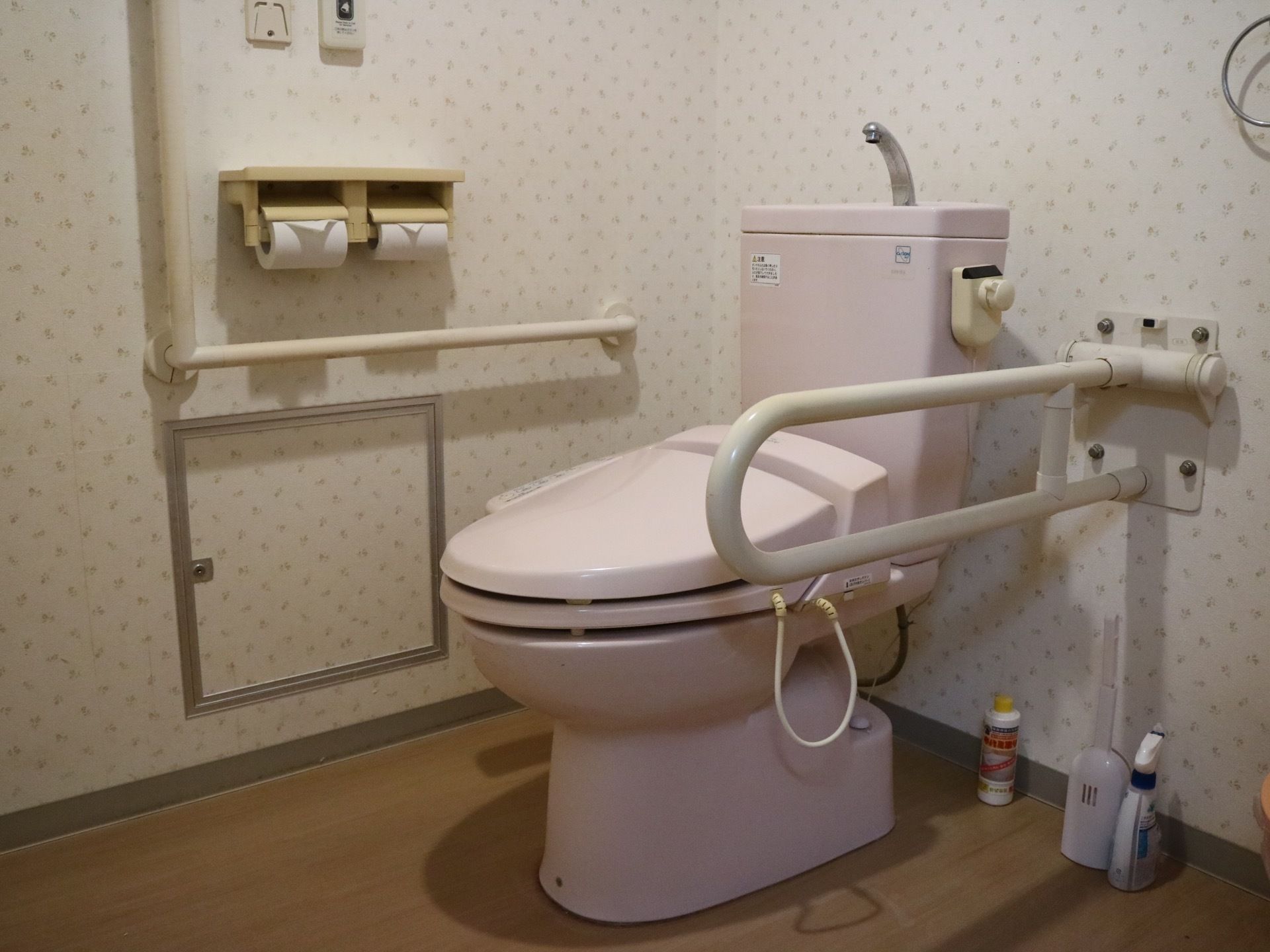 床が木目調で、壁紙が花柄のトイレがある。便座の横や壁には手すりが備え付けられ、ナースコールも設置されている。