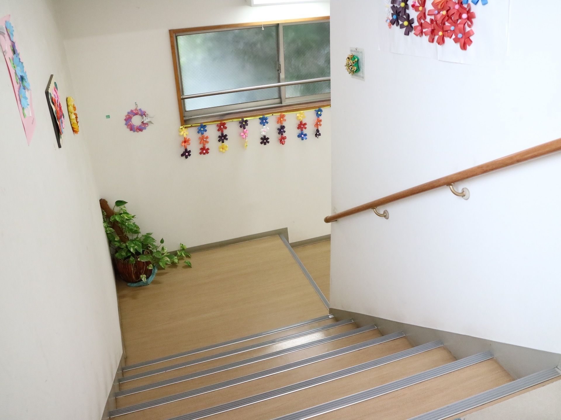 手すりの付いた幅の広い階段がある。踊り場の角には観葉植物が置かれ、壁には色紙で作った花が飾られている。