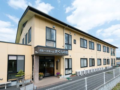 ベージュ系の外壁の２階建ての建物は玄関に屋根があり、ホーム名も描かれている。