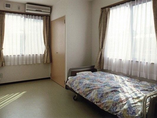 大きな窓が二つあり、それぞれにレースカーテンとカーテンが付き、花柄の寝具がセットされたベッドやエアコン、収納もある。