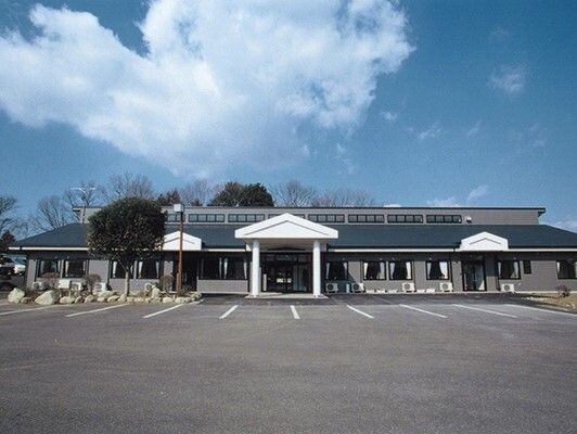 広い駐車場が付いた平屋建ての建物は白とネイビーブルーで、目の前には広い駐車場がある。空には青空が広がっている。