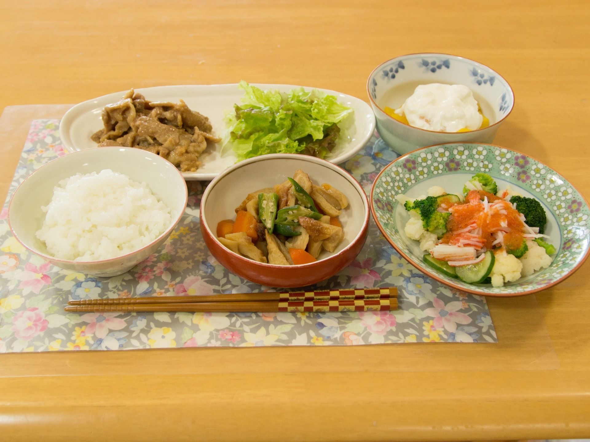 焼肉とレタス、煮物、サラダ、ご飯、フルーツヨーグルトが彩りよく盛られ、ランチョンマットの上に置かれている。