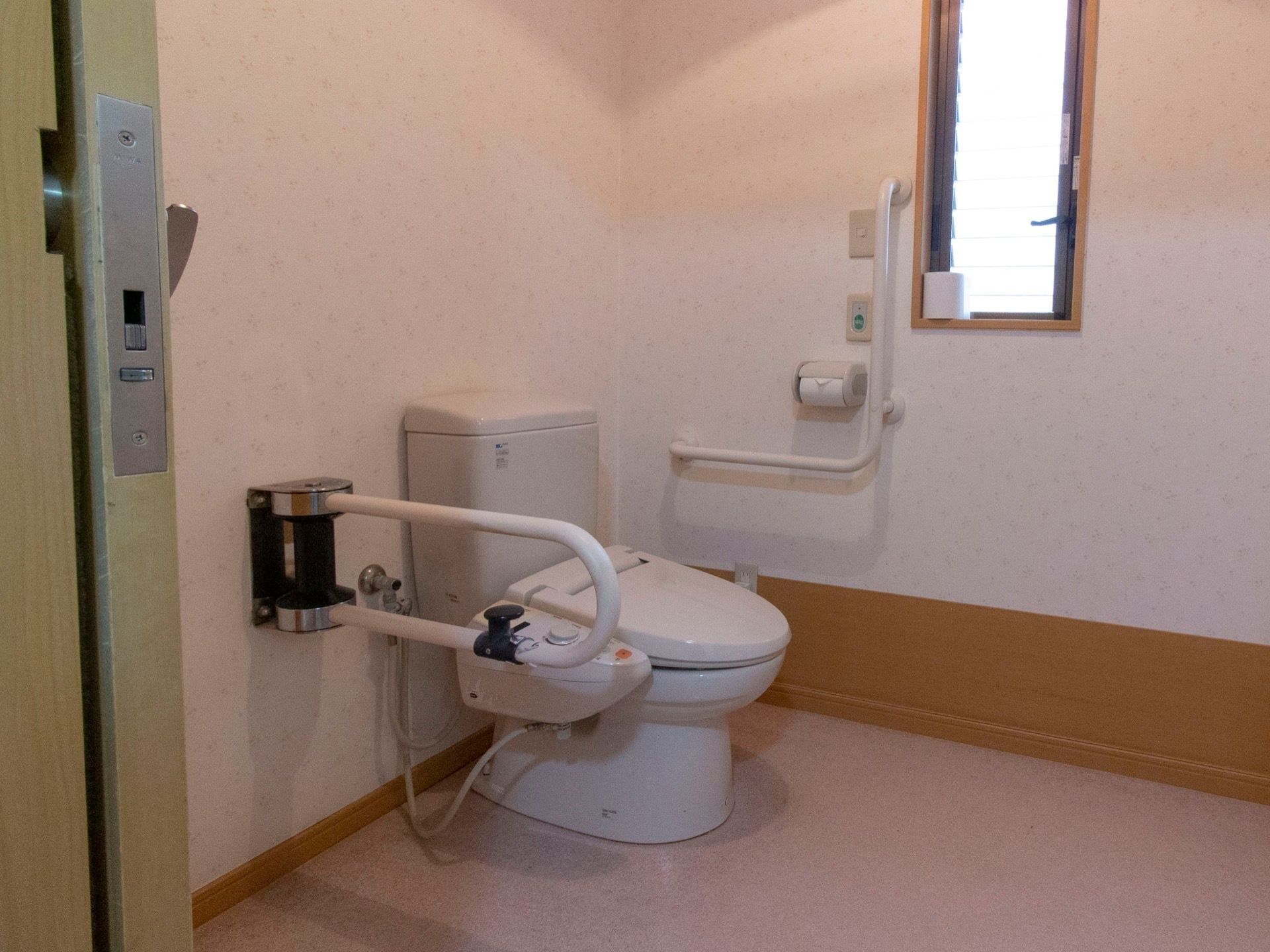 タンク式のトイレの両脇に手すりが設置されている。壁には緊急用の呼び出しボタンがあり、窓には予備のトイレットペーパーが置かれている。