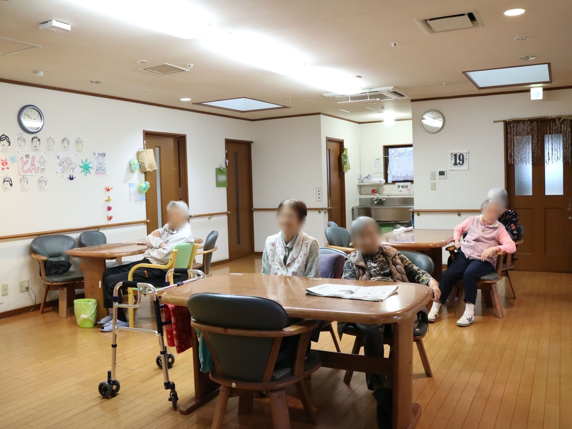 4人掛けのダイニングテーブルと椅子が置かれ、入居者様が座ってくつろいでいる。壁に、似顔絵などが飾られている。
