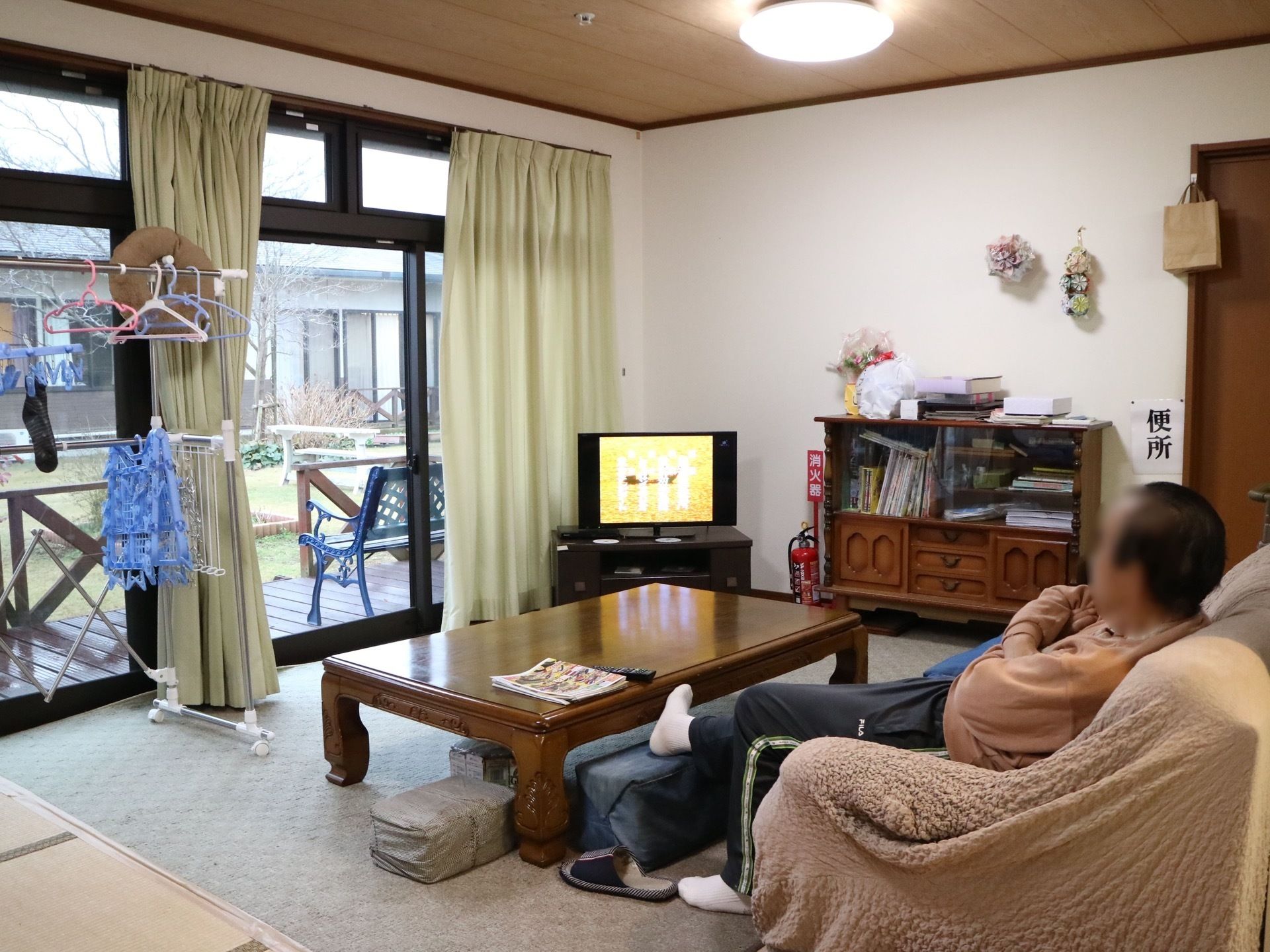 室内の角側にテレビが置かれ、ソファーに座って入居者様がテレビ鑑賞をしている。テレビの横には大きな窓が設置され、ベランダへつながっている。