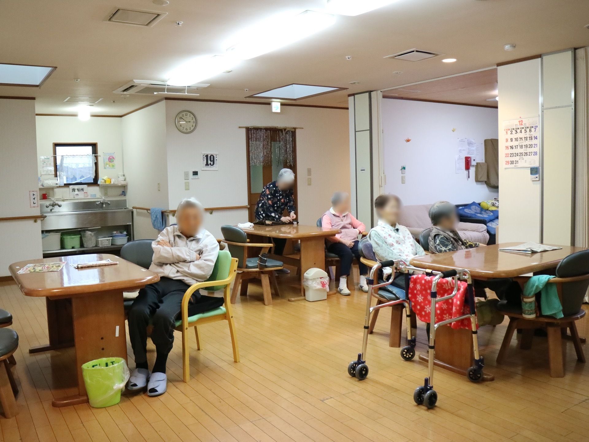 共有スペースにダイニングテーブル、椅子が置かれている。壁に時計やカレンダーがかけられている。奥の方には、洗面台が設置されている。