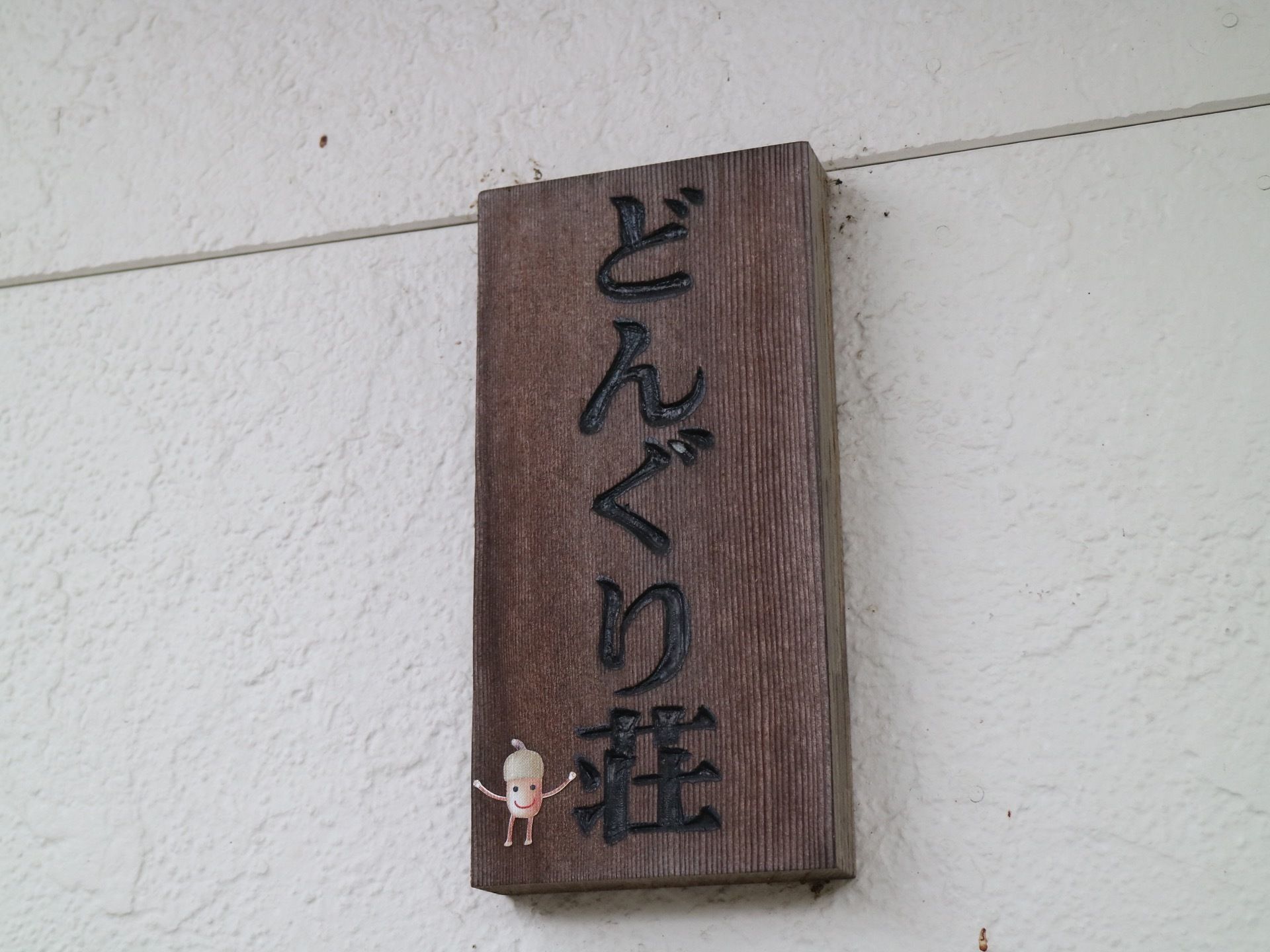 玄関前の外壁は白く、木造の表札が掛けられている。縦向きに施設名が書かれており、どんぐりの絵も描かれている。