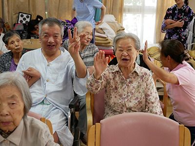 ピンクの椅子がたくさん並べられた共有スペース。白髪の高齢女性と片麻痺の高齢男性が隣同士に座り、笑顔で過ごしている。