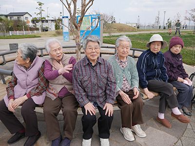 6名の高齢女性が気の周りに設置された円形のベンチに座っている。全員長袖長ズボンで、帽子をかぶっている人もいる。