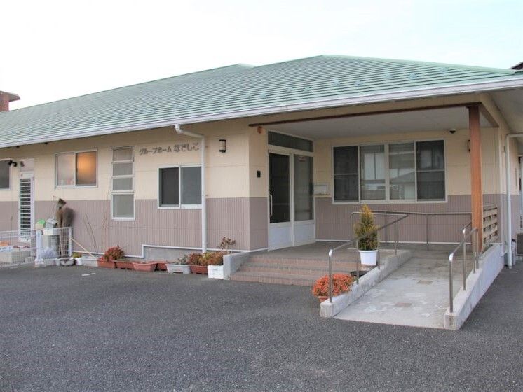 緑の屋根が付いた平屋建ての建物の周りにはプランターが置かれ、玄関には階段と手すり付きのスロープがある。