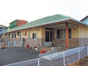 ベージュのジャケットを着た高齢男性に高齢女性が寄り添い、若い女性がそばにいる。3人は広場に座っている。