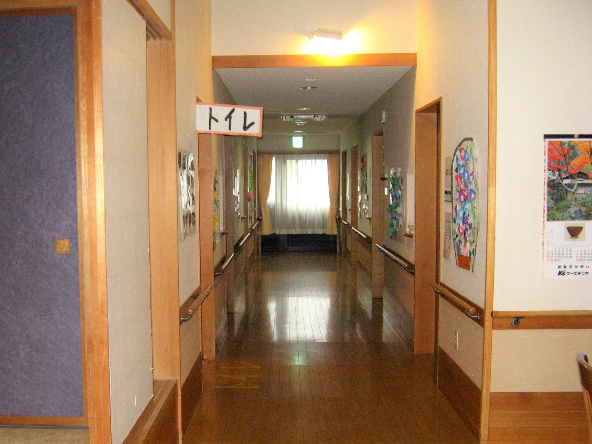 木目調のフローリングの廊下である。突き当たりに非常扉が設置されており、カーテンが取りつけられている。