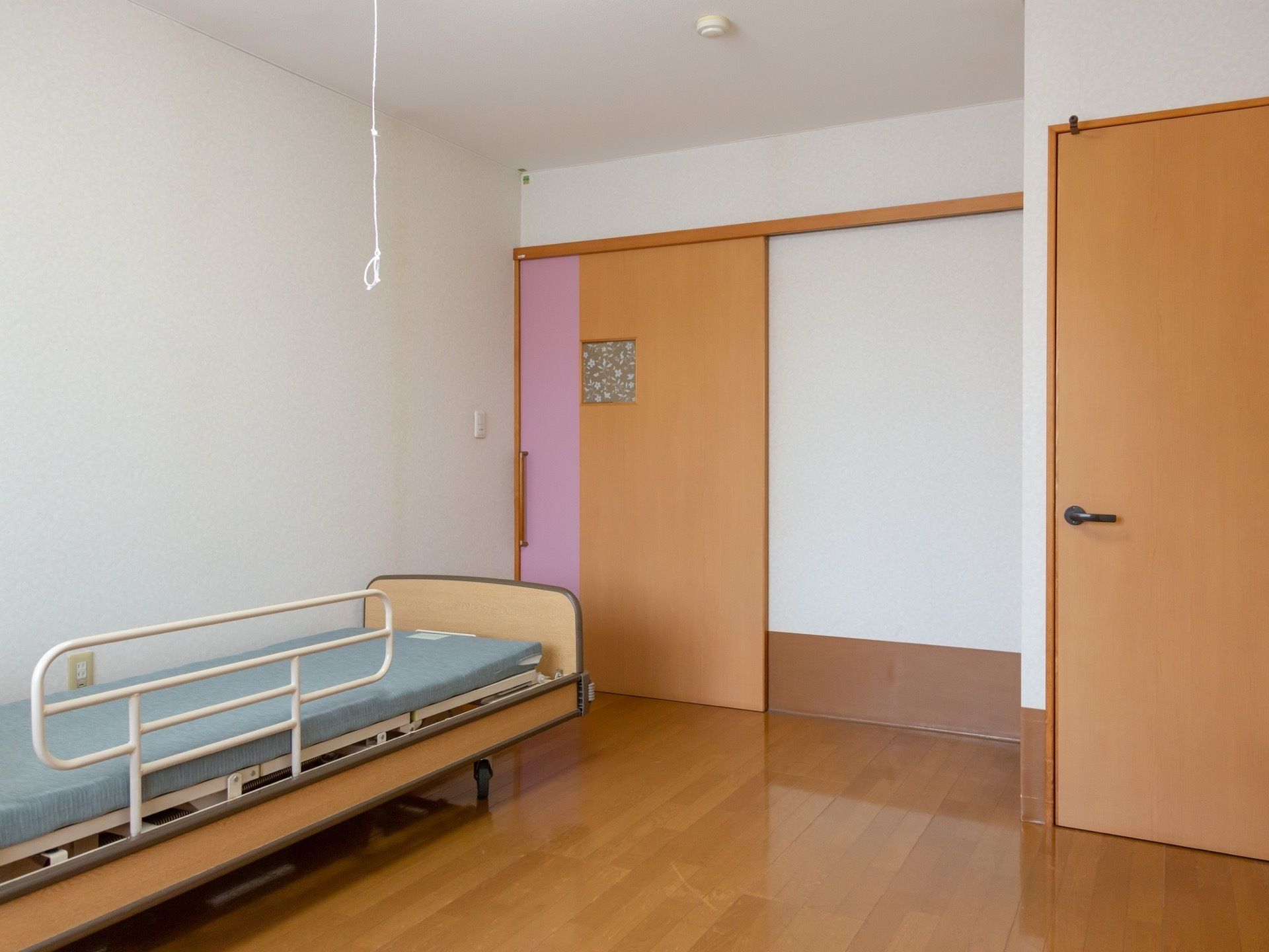 居室内はドアやフローリングなど、あたたかみのあるウッド調でまとめられている。壁際に介護用ベッドが設置されている。