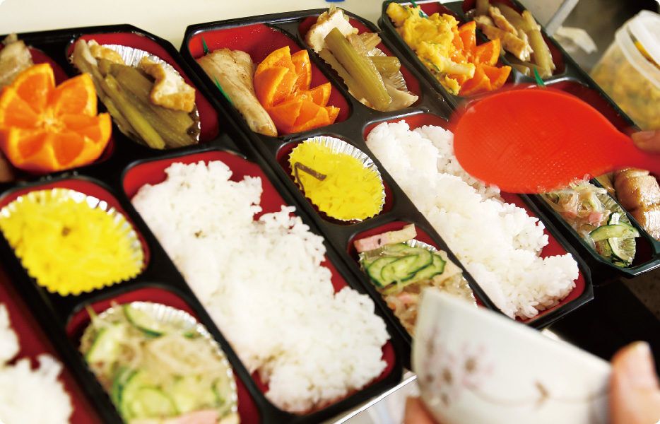 野菜の煮物、春雨サラダ、みかんのデザートが盛り付けられた弁当に、ご飯をつめる様子