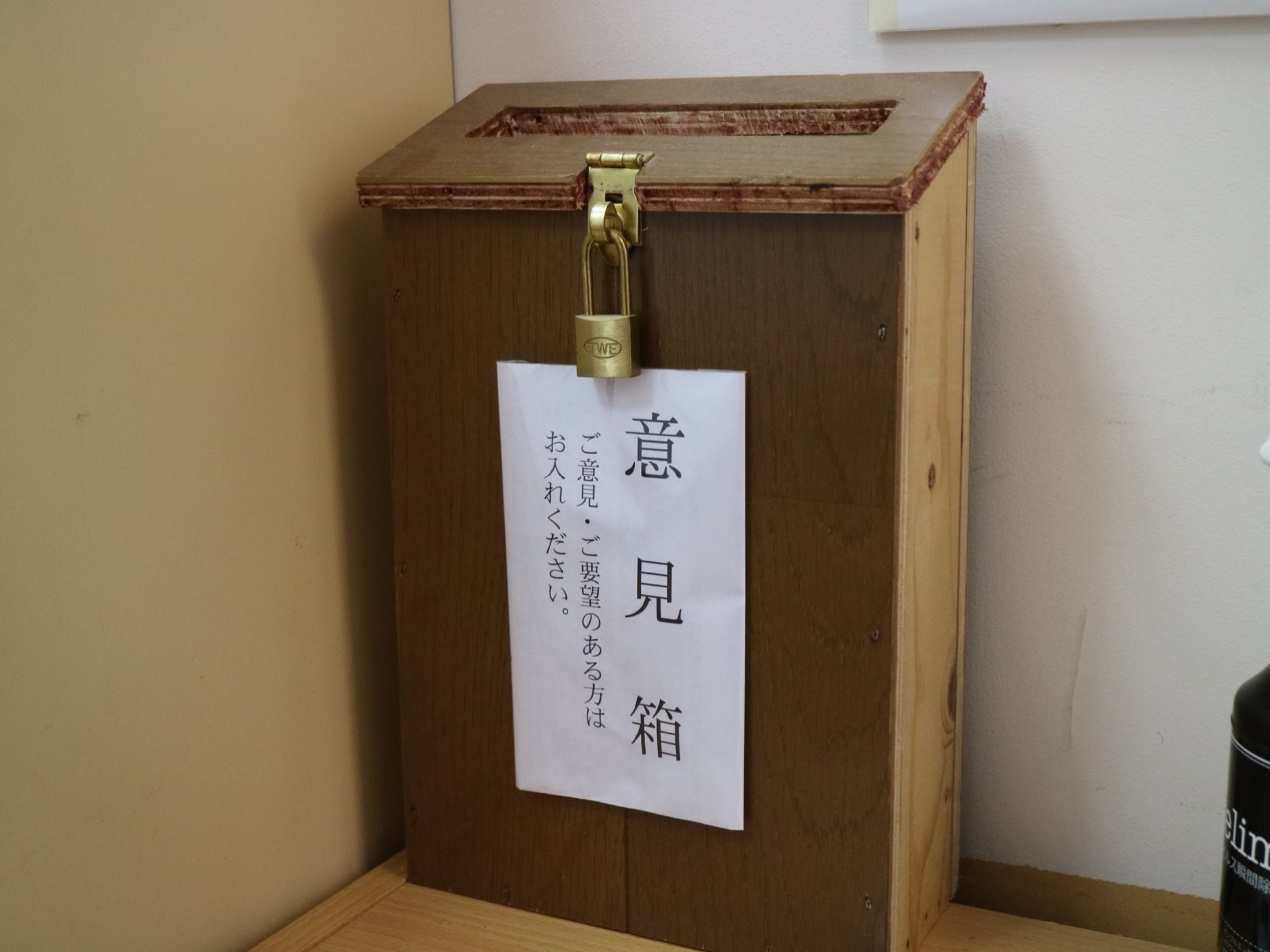木製の棚の上に意見箱が置かれている。茶色い木の箱で作られており、紙で意見箱と表示がされている。錠前がかかっている。