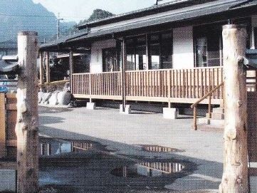 立った樹を途中で切った柱。向こうに平屋で瓦屋根の建物が見える