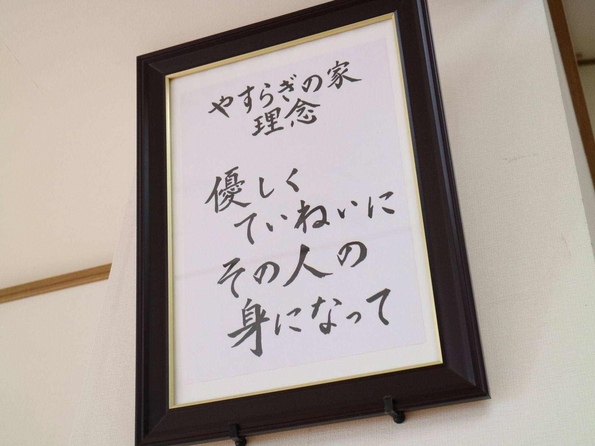 白い壁に黒い枠の額縁が飾られている。施設の理念が習字で書かれていたものが入れられている。額縁はフックに立てかけられている。