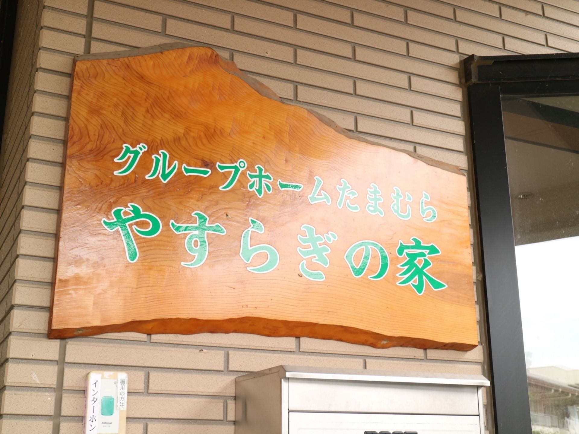 レンガ柄の外壁に板材を使用した看板がかかっている。緑色の字で施設名が表示されている。その下にポストがある。