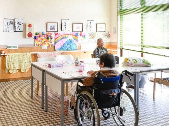 手すりには大判のタオルのようなものが抱えられ、壁には週次などの作品が飾られ、男女の入居者がそれぞれ自分の席についている。