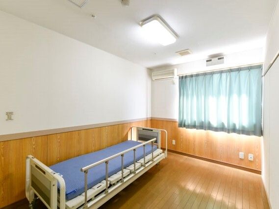 白い壁にフローリングのフロア、壁の下半分ほどは板が貼られている。エアコンや介護ベッドがあり、カーテンとレースカーテンが窓についている。