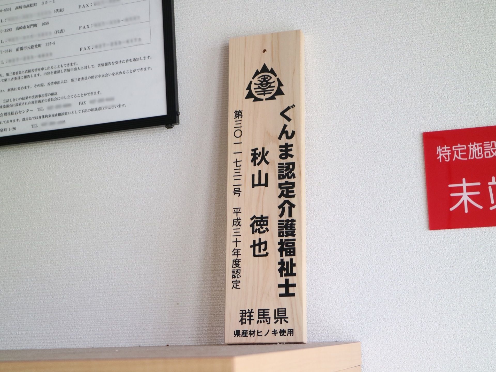 木製の棚の上に小さな板が置かれている。県の認定介護福祉士の名前が書かれている。その左に額縁が貼られている。