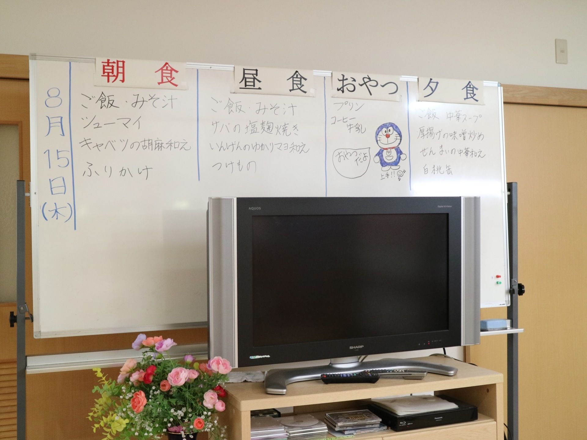 壁際に大きなホワイトボードが置かれ、食事のメニューが書き出されている。その前に大型液晶テレビがある。