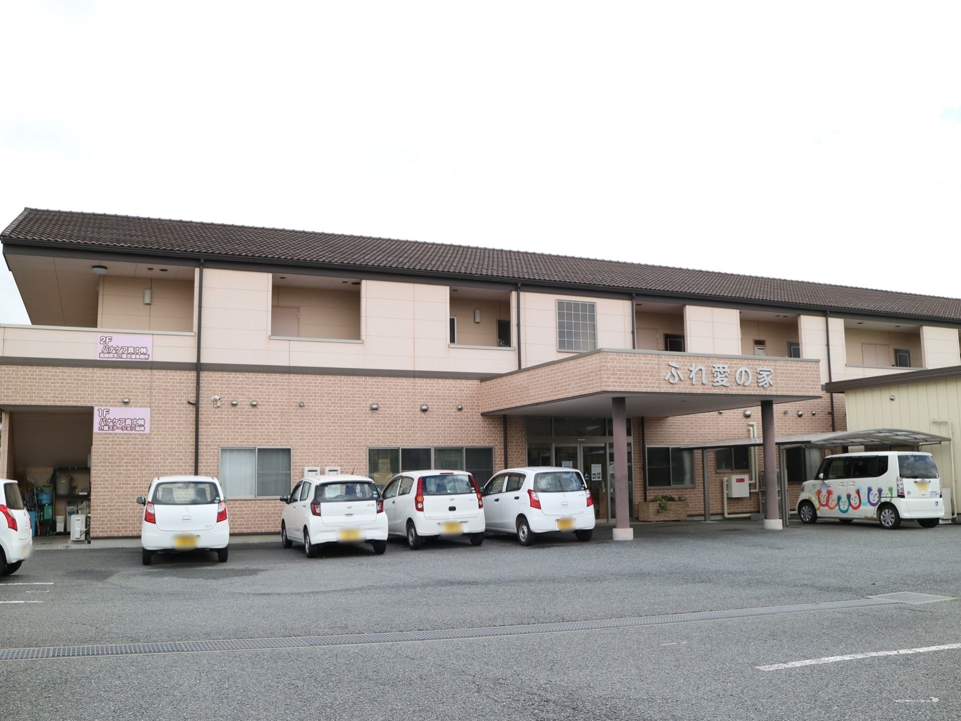 ピンク系統の２色で塗り分けられた外観の建物が建っている。２階建てで中央部分に玄関があり、屋根がせり出している。