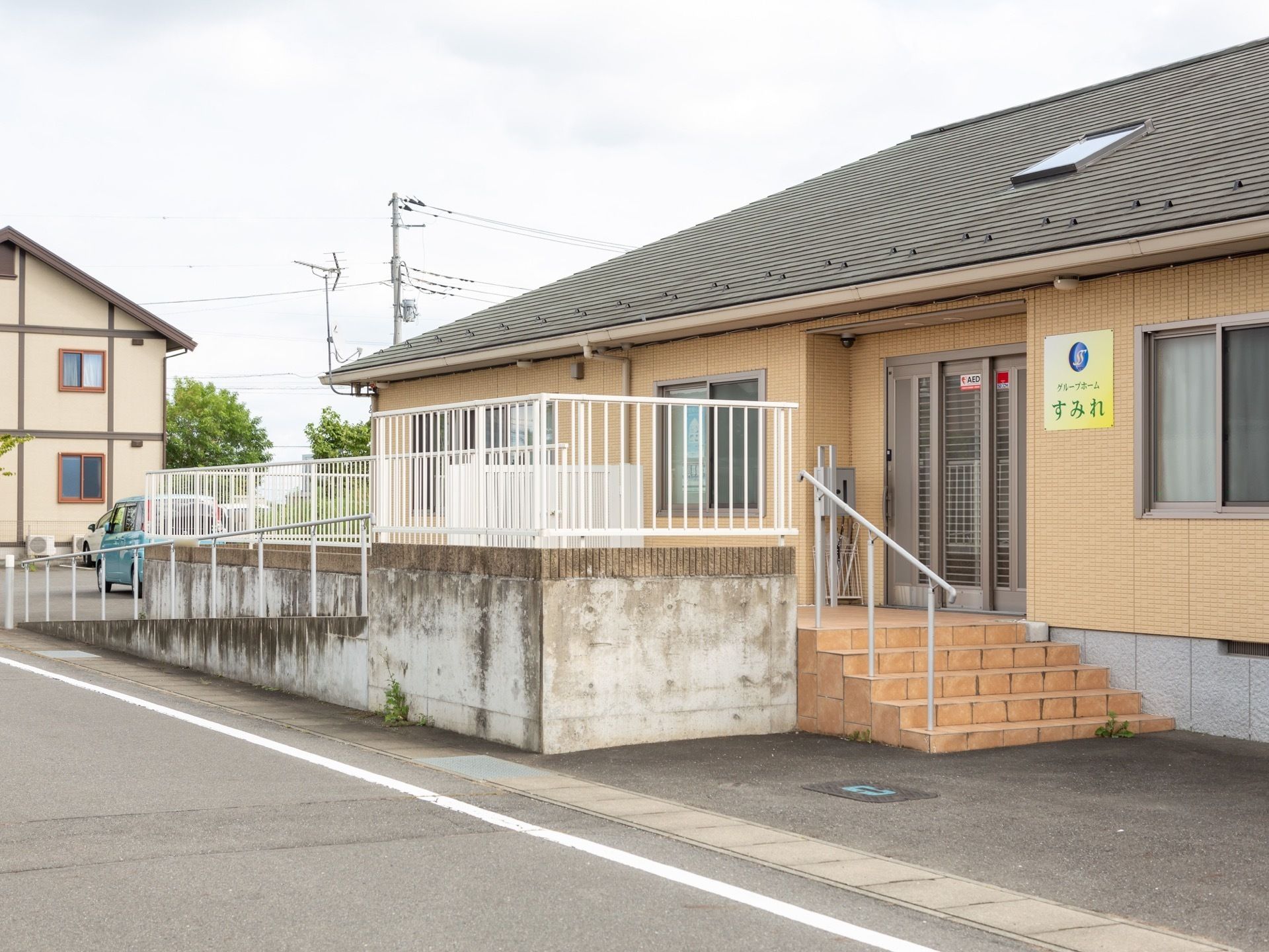 黄色い外壁の平屋建ての建物。玄関の横には施設名が書かれた表札が設置されており、屋根部分に小窓がついている。