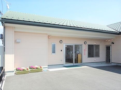 白い壁の平屋建ての建物。グリーンの瓦屋根が付き、建物の玄関の周りには花が植えられたプランターもある。