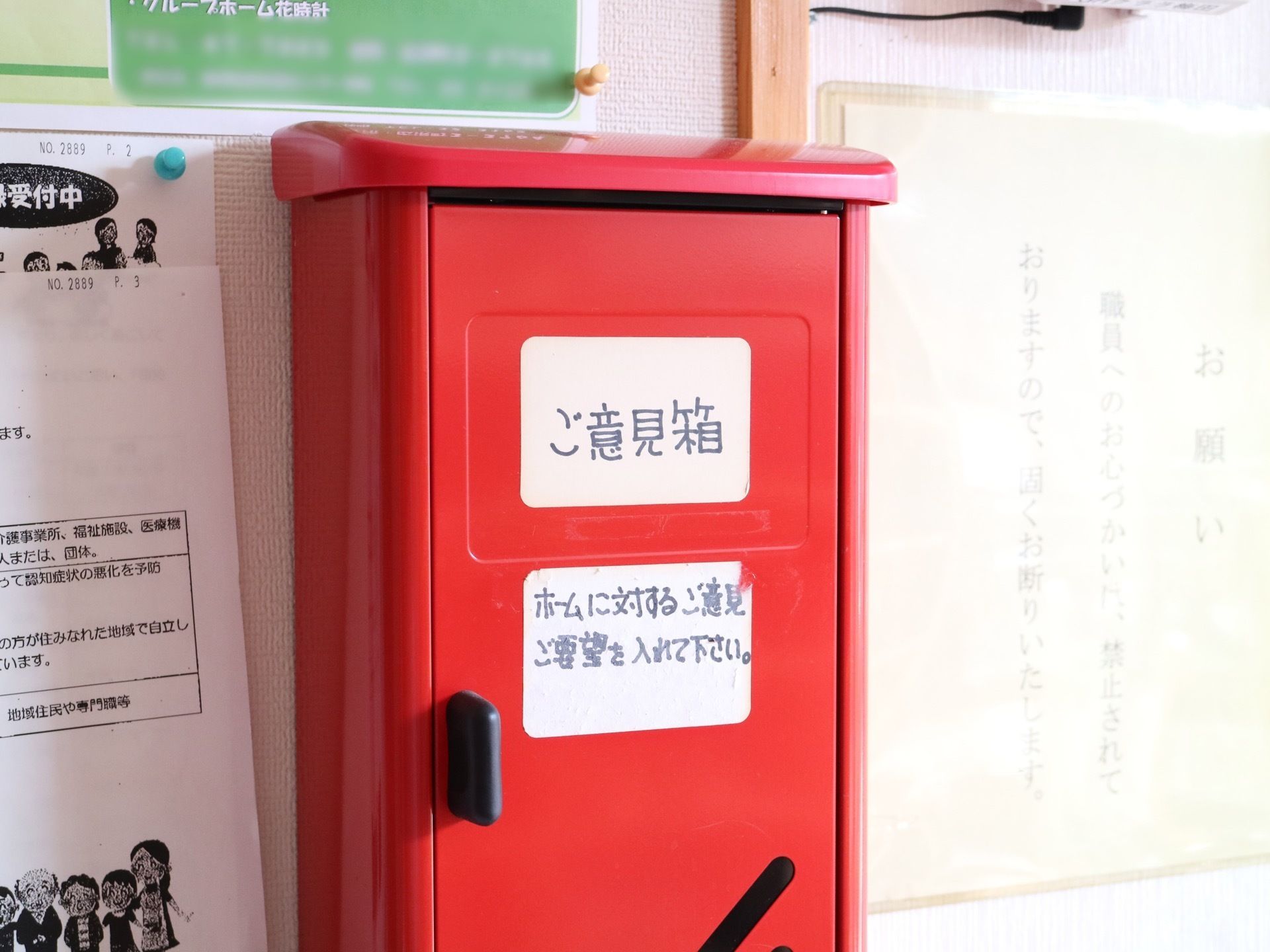 赤いボックスの意見箱が掲示板に取り付けられている。まわりにはチラシやポスターなどが画びょうで留められている。
